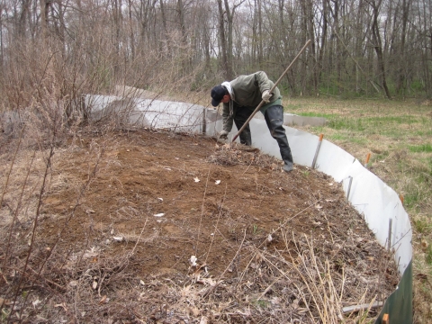 A mound of dirt encompasses the photo, with grassland and forest seen in the background. On the dirt mound, a man with a tool is bended over.