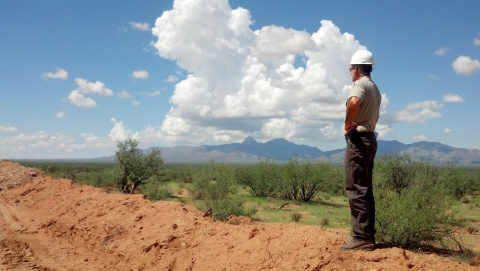 A man in a hard hat stands on a mound of dirt and looks out over patchy grass and tall bushes with mountains in the background.