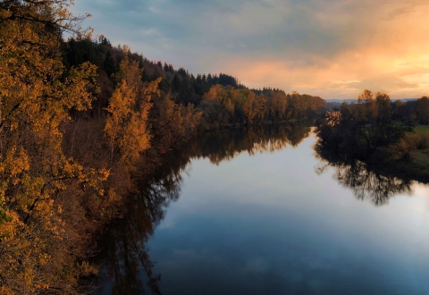 A glowing sky is reflected in Lake River that has fall vegetation along it