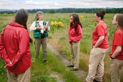 A woman speaks to group of people in an open field with a tree line in the distant background.