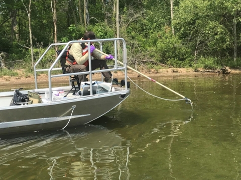 A biologist seated on a boat holds a long pole in the water. A tube attached to the pole is connected to a water collection box on the boat. 
