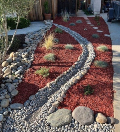 Small rounds of shaggy grass pop up through bark. Plants are on either side of a manmade water channel that winds through a front yard.