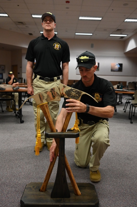 Two honor guard employees set golden Pulaskis in a holder. One is kneeling and one is standing at attention