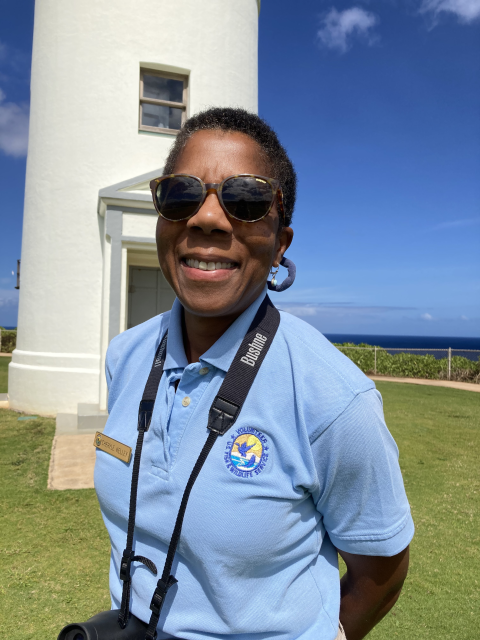 A woman with sunglasses and binoculars smiles in front of a lighthouse. She is wearing a light blue uniform.