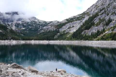 A view of a blue lake surrounded by mountains and trees. At the base of the lakes stands a person, small in scale to the surrounding landscape