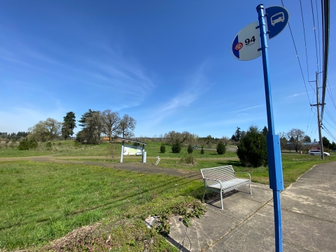 Image of a blue pole with a metal flag on top with a bus symbol and the number 94. In the background is a silver metal bench, grass, a gravel trail and an map kiosk.