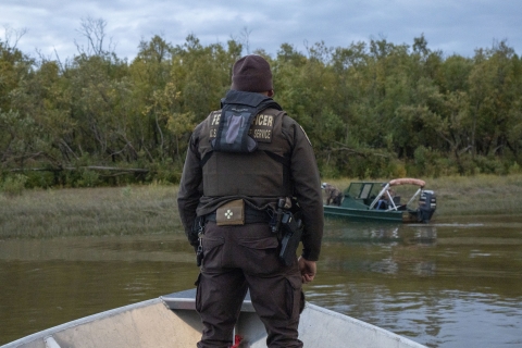 A uniformed Federal Wildlife Officer stands on the bow of a boat as it nears a hunters boat on the shoreline of a river. Thick woodland on the shore of a river. 