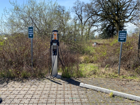 Image of an electric car charging station in the middle with two vertical signs on either side that read hybrid vehicle parking only with trees in the background.