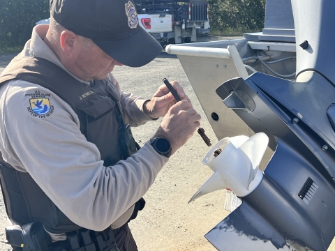 A uniformed officer works on a boat propellor that is on an elevated motor mounted on a boat that is on a trailer in a parking lot. 