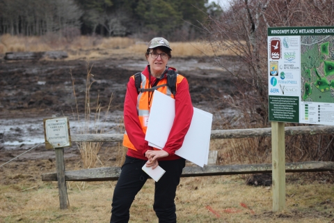 a woman wearing glasses, a green ball cap, red jacket, and orange vest and holding large white papers under her arm stands before an area of mud 