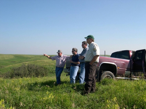 Four people stand in a grassy, green field. Three men are dressed in jeans and short-sleeved shirts. One man is wearing a U.S. Fish and Wildlife Service uniform. They survey the land together.