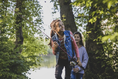 Two kids holding fishing rods walk through the trees near New England lake. 