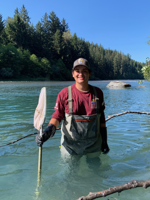 AmeriCorps Service Member in waders, standing in a river while holding a net. Trees and blue sky in the background.