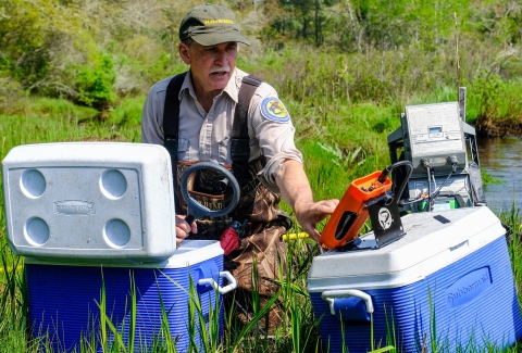 a white man wearing a green ball cap, tan shirt, and brown camouflage waders adjusts a setting on an orange box resting on a blue and white cooler