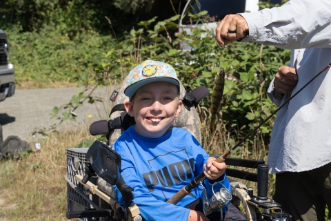 A smiling boy wearing a blue shirt and a patterned cap sits in a motorized wheelchair, holding a fishing rod while an adult beside him lifts his freshly caught fish, still on the fishing line. Green blackberry bushes are visible in the background.