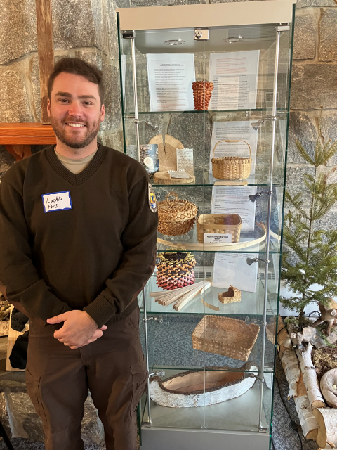 Man in refuge browns smiles in front of display case full of weaved baskets.