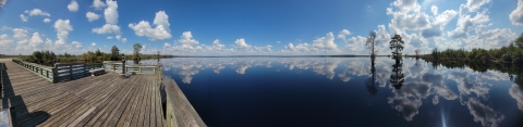 A deck on a lake. Trees and clouds are reflected on the lake.