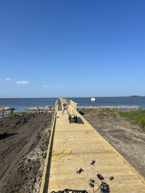 A pier with fresh wood decking extends past a gravel parking lot, a marshy shoreline, and a rocky breakwater into a wide-open body of water. Various construction tools and workbenches are set up at the start of the pier.