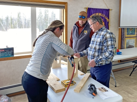 Two people using a nail gun to construct a wooden box with one person watching in the background. The work occurs indoors on a white table that has some wood pieces on it.