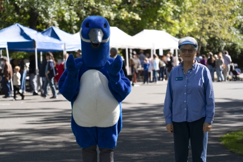 Puddles the blue goose mascot and a volunteer stand in front of a bunch of tents and people.