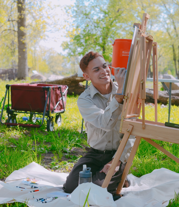 Logan Sauer paints on an easel out in nature on a sunny day.