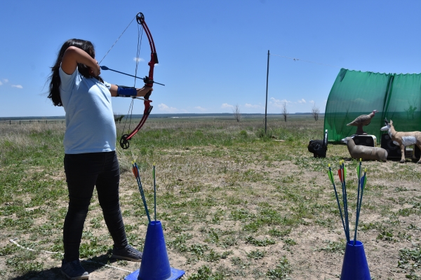 A young girl pulling back on a bow with a notched arrow aiming at an archery target.