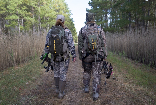 Two hunters in camouflage holding compound bows walk away from the camera down a wide path through a pine forest.