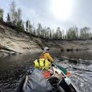 Tim Ericson, sitting at the front of a boat, paddles on the Deshka River. The photo is taken from the back of the boat, and Ericson looks back at the camera over his shoulder. The boat is filled with supplies, including moose antlers.