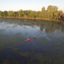 Aerial view looking down on two kayakers in a marsh with lots of green aquatic vegetation on surface, green forest in background