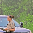 Two law enforcement officers -- a man and a woman -- speaking as the male officer points. They are in front of a police car in a wooded setting.