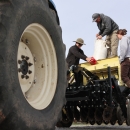 Three people standing on a tractor-pulled seed planter, with a large tractor tire in the foreground.