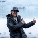 A young woman in a dark rain jacket smiles and holds up two thumbs up. She stands in front of icy water, with a large glacier visible in the background.
