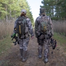 Two hunters in camouflage holding compound bows walk away from the camera down a wide path through a pine forest.
