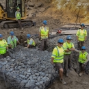 Ten people wearing bring yellow shirts and blue hard hats pose near a newly constructed rock wall