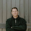 David Viker stands arm-crossed in a black jacket with blue jeans in front of wooden fence atop of a grey brick wall.