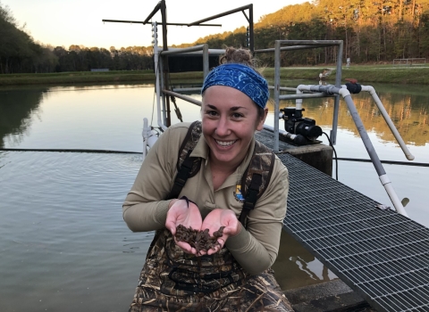 Jessica Radich holds mussels while wearing waders and standing in shallow water in front of a dock.
