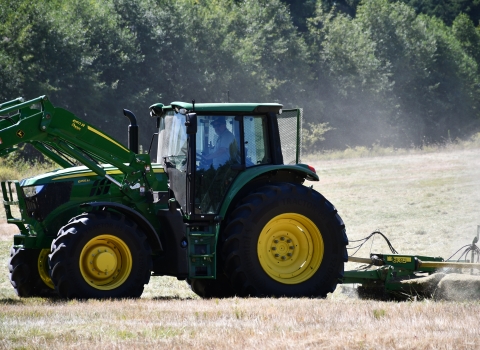A green and yellow tractor tows a mowing attachment across a dusty field of grass with evergreens in the background.
