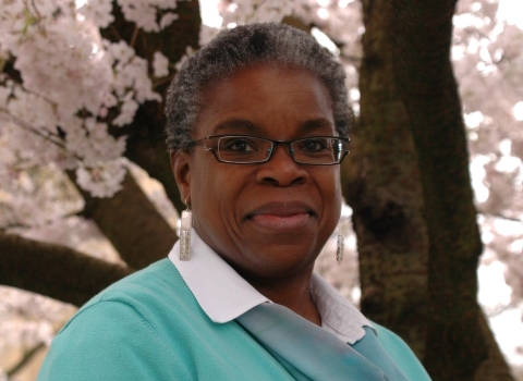 A woman wearing glasses and a blue sweater smiles while standing under a blooming cherry blossom tree