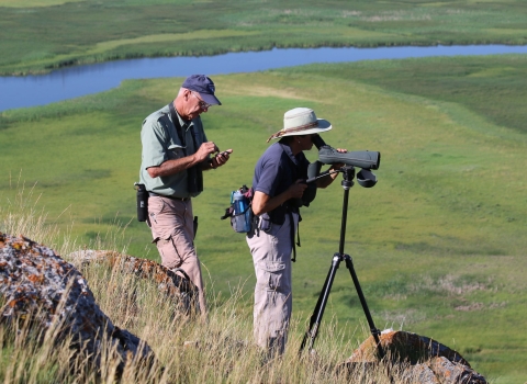 Volunteers Chuck and Betty Mulcahy, wearing hats and hiking clothes, survey trumpeter swans and cygnets weekly from atop a butte at National Elk Refuge in Wyoming.