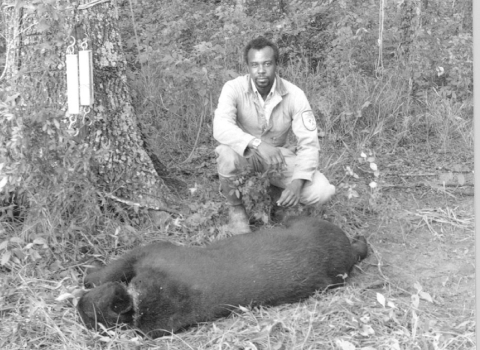 A black and white portrait of a black man -- A Fish and Wildlife Service employee, squatting behind a sedated, trapped bear