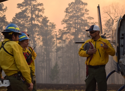 Four firefighters stand next to a helicopter talking. The background is smoky with large trees.