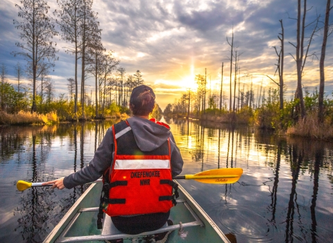 woman in front of canoe holding a yellow paddle in dark reflective water with cypress trees and clouds in a sunny sky. 