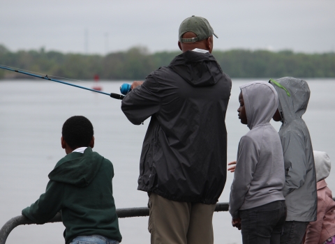A man stands on the waterfront with a fishing rod surrounded by kids