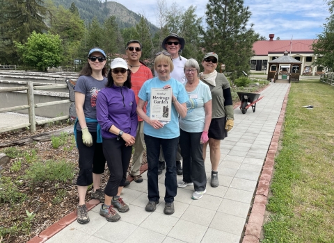A group of 7 people stand in front of the pollinator garden holding a heritage garden sign