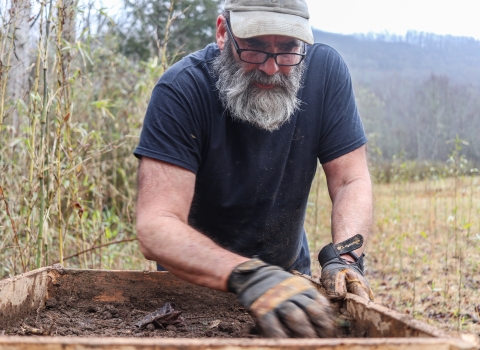 Man standing on the edge of a field, wearing work gloves, and sorting through dirt in a large sieve in front of him